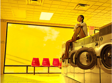 Charger l'image dans la galerie, Une femme assise sur des machines à laver. Pièce jaune vif, chaises rouges, ciel jaune.