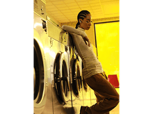 Charger l'image dans la galerie, Une femme s'appuie sur des machines à laver, porte des écouteurs, avec un fond jaune.