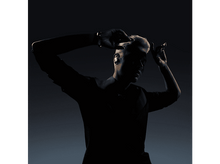 Charger l'image dans la galerie, Silhouette d'une personne aux cheveux blonds, portant des écouteurs, mains en l'air, fond sombre.