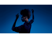 Charger l'image dans la galerie, Une femme avec un casque, les bras levés, de profil sur un fond bleu, vêtue de vêtements sombres.