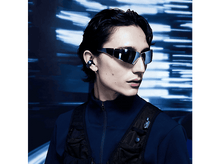 Charger l'image dans la galerie, Jeune homme portant des lunettes de soleil, un gilet noir et un écouteur. Fond bleu. Aspect moderne.