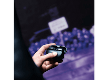 Charger l'image dans la galerie, Main tenant des écouteurs noirs dans un étui de chargement ouvert, avec un arrière-plan flou d'un magasin.