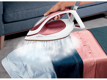 Charger l'image dans la galerie, Une main repassant une chemise avec un fer à repasser rose et blanc, vapeur visible, avec des tissus bleus et roses.