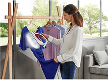 Charger l'image dans la galerie, Femme repassant une robe bleue. Portant à vêtements en arrière-plan. Fenêtre et canapé.