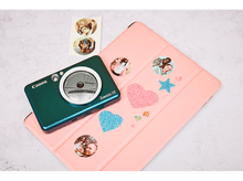 Charger l'image dans la galerie, Un appareil photo Canon Zoemini S2 vert, une tablette rose et des autocollants. Les articles sont sur une surface blanche.