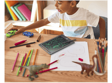 Charger l'image dans la galerie, Garçon dessinant à une table avec des jouets, des crayons et une tablette de dessin. Arrière-plan flou.