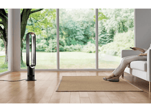 Charger l'image dans la galerie, Une femme assise sur un canapé blanc, tenant une télécommande. Un ventilateur noir à côté. Grande fenêtre en arrière-plan.