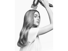 Charger l'image dans la galerie, Une femme aux longs cheveux utilise un sèche-cheveux. Le sèche-cheveux est tenu au-dessus de sa tête. Elle porte un haut blanc.