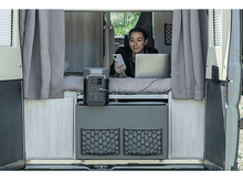 Charger l'image dans la galerie, Femme dans un van utilisant un téléphone et un ordinateur portable. Une station d'alimentation portable est sur un meuble gris, et des rideaux sont suspendus.