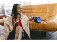 Charger l'image dans la galerie, Femme avec tresses près canapé jaune, bouteille bleue. Veste beige, haut rouge.