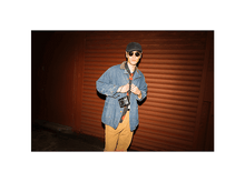 Charger l'image dans la galerie, Une personne en veste en jean et chapeau se tient devant une porte à volet rouge, tenant un appareil photo.