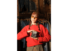 Charger l'image dans la galerie, Une personne en pull rouge tient un appareil photo Instax Mini 41. Arrière-plan : magasin de vêtements.