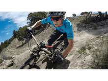 Charger l'image dans la galerie, Un cycliste sur un VTT descend un sentier escarpé et sale. Portant un casque, des lunettes de soleil et des gants.