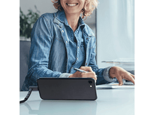 Charger l'image dans la galerie, Une femme sourit en travaillant à un bureau. Un téléphone noir est devant elle.