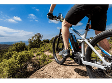 Charger l'image dans la galerie, Personne faisant du vélo électrique sur un sentier rocheux sous un ciel bleu avec des arbres.
