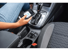 Charger l'image dans la galerie, Une personne dans une voiture tient un téléphone affichant une application de cartographie. Le levier de vitesse et la console sont visibles.