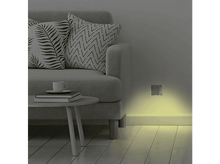 Charger l'image dans la galerie, Un canapé avec coussins, une table d'appoint et une prise murale avec lumière jaune.