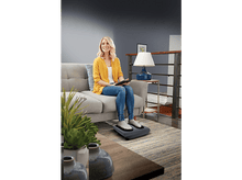 Charger l'image dans la galerie, Femme assise sur un canapé gris utilisant un masseur de pieds, avec une table d'appoint et une lampe. Fond neutre.