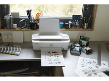 Charger l'image dans la galerie, Une imprimante blanche imprimant un papier avec un motif floral, à côté de cartouches d'encre et de fournitures de bureau.