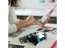 Charger l'image dans la galerie, Une femme regarde des papiers imprimés et des cartouches d'encre sur une table. Les cartouches sont de la marque HP.