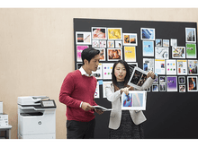 Charger l'image dans la galerie, Deux personnes discutent d'images devant un mood board. L'un tient des photos, l'autre un papier. Bureau.