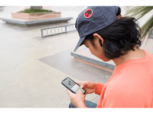 Charger l'image dans la galerie, Homme portant une casquette et des écouteurs, regardant un téléphone. Arrière-plan flou d'un skatepark.