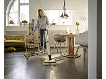 Charger l'image dans la galerie, Une femme nettoie un sol gris avec un balai électrique jaune et blanc dans une pièce lumineuse.