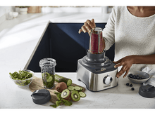 Charger l'image dans la galerie, Femme utilisant un mixeur pour faire un smoothie, ingrédients sur la table. Cadre de cuisine.