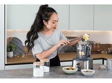 Charger l'image dans la galerie, Femme ajoutant des ingrédients à un robot culinaire. D'autres ingrédients sont visibles.