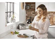 Charger l'image dans la galerie, Femme dans une cuisine tenant un récipient de mixeur. Il y a un mixeur et des ingrédients frais sur le comptoir.