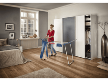 Charger l'image dans la galerie, Une femme accrochant un jean sur un étendoir dans une chambre lumineuse.