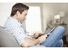 Charger l'image dans la galerie, Un homme utilise une tablette assis sur un canapé. Il porte une chemise à carreaux et un jean. Un arrière-plan flou.