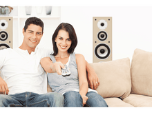Charger l'image dans la galerie, Un couple sourit sur un canapé. La femme pointe une télécommande. Des enceintes sont en arrière-plan.