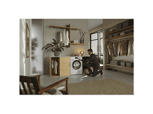 Charger l'image dans la galerie, Un homme s'agenouille près d'une machine à laver dans une buanderie moderne. Des vêtements sont suspendus sur des étagères. Une plante et une chaise en bois sont visibles.