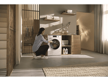 Charger l'image dans la galerie, Une femme utilise une machine à laver blanche. La porte de la machine est fermée. Le fond est une pièce.