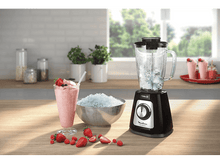 Charger l'image dans la galerie, Un blender, un smoothie et de la glace pilée sont posés sur une surface en bois. Des fraises et des framboises sont éparpillées. Des plantes vertes et une fenêtre sont en arrière-plan.