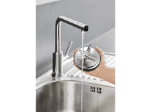 Charger l'image dans la galerie, Un évier avec un robinet, l'eau coule. Une personne tient une pièce métallique de robot culinaire sous l'eau.