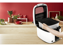 Charger l'image dans la galerie, Mains plaçant un récipient dans un appareil de cuisine blanc et noir, avec un fond de mur rouge.