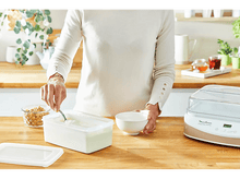 Charger l'image dans la galerie, Une personne préparant du yaourt, avec un bol et un récipient, sur un comptoir en bois.