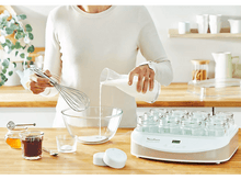 Charger l'image dans la galerie, Femme versant du lait dans un bol, avec un fouet, des pots et une yaourtière.