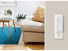 Charger l'image dans la galerie, Un répéteur WiFi Netgear branché sur une prise murale, avec un ordinateur portable et un canapé en arrière-plan.
