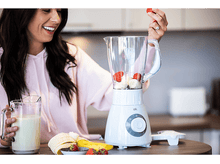 Charger l'image dans la galerie, Femme ajoutant une fraise dans un blender avec des fruits et un pot de lait.