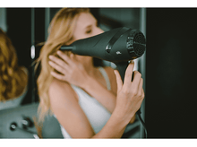 Charger l'image dans la galerie, Femme blonde utilisant un sèche-cheveux noir. Miroir et mur sombre en arrière-plan.