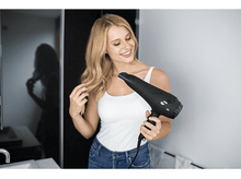 Charger l'image dans la galerie, Une femme sourit en tenant un sèche-cheveux noir. Elle est dans une salle de bain.