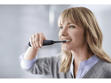 Charger l'image dans la galerie, Femme se brossant les dents avec une brosse à dents électrique bleue et noire, souriante, regardant à droite.
