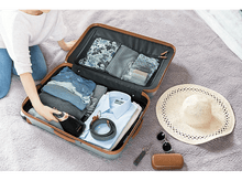 Charger l'image dans la galerie, Une femme prépare une valise avec des vêtements, des accessoires et un chapeau.