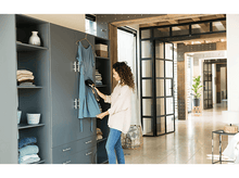 Charger l'image dans la galerie, Femme repassant une robe bleue à l'intérieur d'une armoire grise, avec la lumière d'une fenêtre.