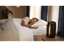 Charger l'image dans la galerie, Une femme dormant dans son lit à côté d'un humidificateur noir sur une table de chevet.
