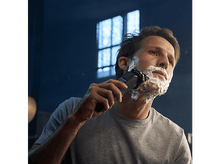 Charger l'image dans la galerie, Un homme se rase avec un rasoir électrique argenté et noir, avec de la crème à raser sur le visage.