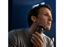 Charger l'image dans la galerie, Un homme se rase le visage avec un rasoir électrique. Fond sombre.